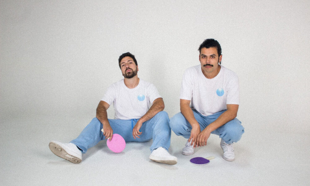 Fotograma del videoclip “Fotos” de Akinorite. El dúo chileno aparece en un set blanco iluminado con luces de neón, acompañado por patinadoras que se mueven alrededor, en una puesta en escena minimalista y retrofuturista.