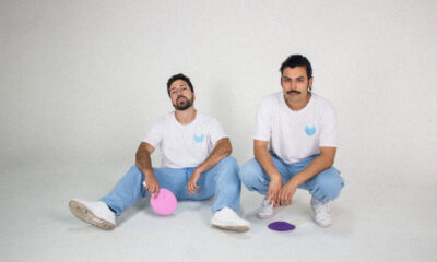 Fotograma del videoclip “Fotos” de Akinorite. El dúo chileno aparece en un set blanco iluminado con luces de neón, acompañado por patinadoras que se mueven alrededor, en una puesta en escena minimalista y retrofuturista.